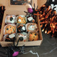 Box of Halloween-themed cupcakes / oat balls with decorative pumpkins and tinsel on a dark surface.